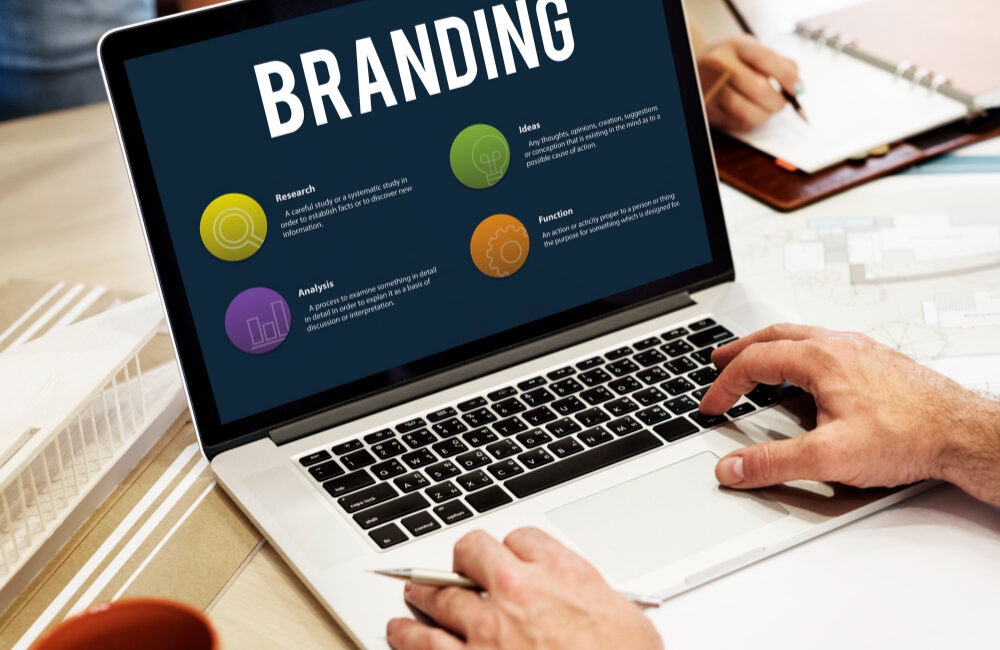 A person working on a laptop displaying a presentation slide titled “Branding” with colorful icons and sections labeled Research, Ideas, Analysis, and Function. A notebook, pen, coffee mug, and architectural model are also on the desk.