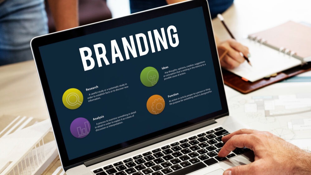 A person working on a laptop displaying a presentation slide titled “Branding” with colorful icons and sections labeled Research, Ideas, Analysis, and Function. A notebook, pen, coffee mug, and architectural model are also on the desk.