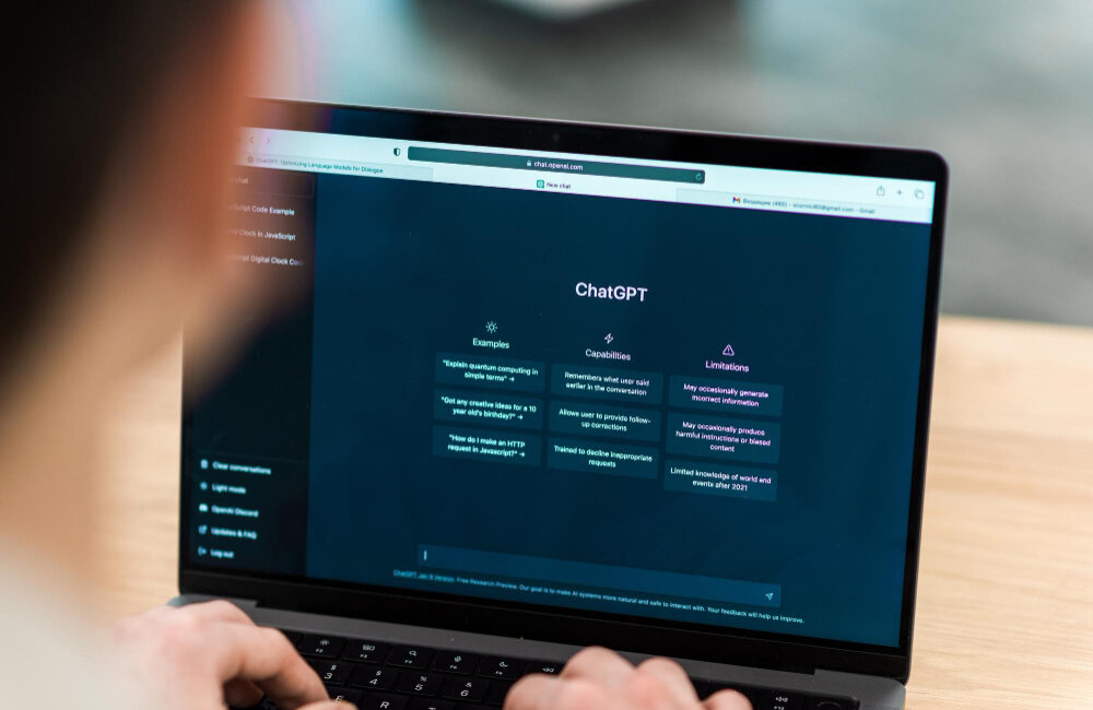 A person using a laptop with the ChatGPT interface open on the screen, displaying examples, capabilities, and limitations of the AI tool.