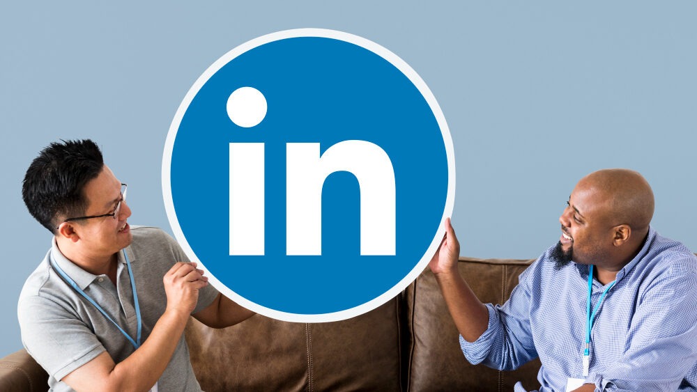 Two people sitting on a brown couch smiling and holding a large round LinkedIn logo together against a light blue background.